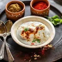 Dahi vada in a white ceramic bowl garnished with chilli powder and coriander on a dark rustic platter